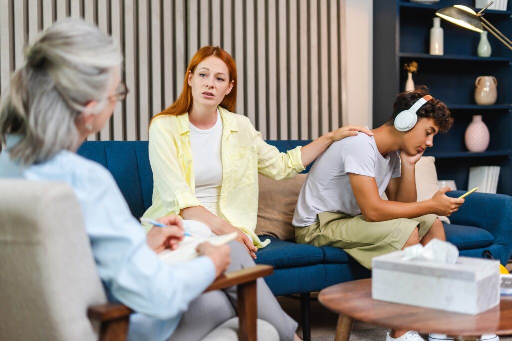 Mother comforting sad teenage son during therapy session with psychologist, teenager using earphones